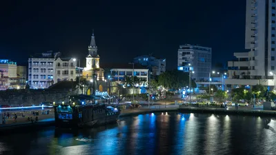 City waterfront at night with illuminated buildings and a boat reflecting in the water.