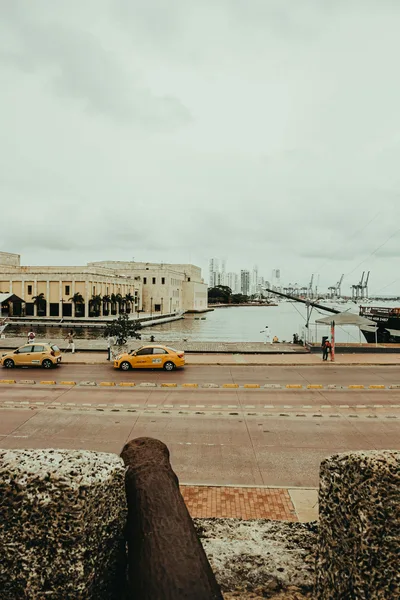 View of a historic building and waterfront with yellow taxis in the foreground, cloudy sky above.